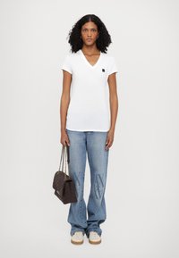 Camiseta blanca de algodón con escote en V, de manga corta, combinada con unos jeans azul claro con detalles estampados. La modelo sostiene un bolso marrón acolchado.