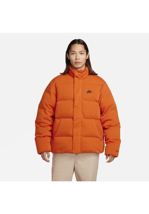 Orange puffer jacket with a high collar, square quilted design, front pockets, and black logo accent on the chest. Soft, insulated material.