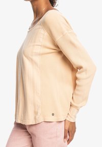 Chemise beige en maille gaufrée à manches longues avec un col rond et des coutures latérales. Présente un petit logo à l'ourlet et un tissu texturé.