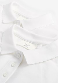 Weiße Baumwoll-Schulpoloshirts mit gewellten Kragen und Dreiknopfleisten, gestapelt mit sichtbarem "next school"-Etikett im Kragen.
