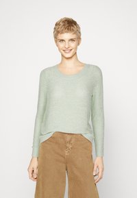 Maglione lavorato a maglia verde chiaro con maniche lunghe, scollatura rotonda e polsini a costine; indossato con pantaloni larghi color sabbia.