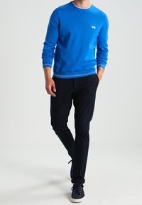 Niebieski sweter z okrągłym dekoltem, z białymi wykończeniami przy mankietach i dolnym brzegu, w połączeniu z czarnymi spodniami i niebieskimi trampkami. Prosty design, luźny krój.