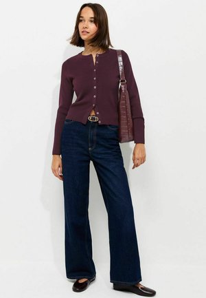 Femme portant un cardigan violet boutonné, un jean large foncé, des chaussures noires et un sac à main violet assorti.