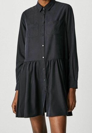 Frau trägt ein langärmeliges schwarzes Hemdkleid mit zwei Brusttaschen und einem gerafften Rock, steht vor einem schlichten Hintergrund.