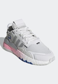 Białe sportowe buty z teksturowaną siateczką na cholewce, szarymi akcentami, różowo-niebieską podeszwą oraz płaskimi sznurowadłami. Widoczne trzy paski po bokach.