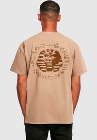 Beige t-shirt met korte mouwen met een grafisch ontwerp van een farao omgeven door hiërogliefen, gedrukt in bruin op de achterkant.