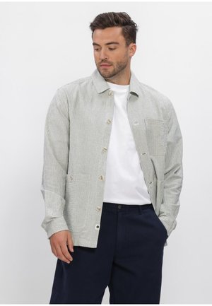 Man draagt lichtgrijze gestreepte jas over wit overhemd en donkerblauwe broek, staat met één hand in de zak en kijkt naar beneden links.