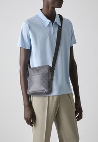 Bolso bandolera gris con un diseño elegante y correas ajustables, llevado sobre una camisa polo azul claro y pantalones beige.