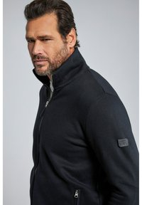 Chaqueta de forro polar negra con cremallera en un diseño ajustado, que presenta un cuello alto y bolsillos laterales. Incluye un pequeño parche con el logo en la manga.