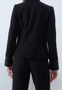 Černý přizpůsobený blazer se strukturovanými rameny, vyrobený z hladké látky, s elegantními liniemi a koženými akcenty na manžetách.