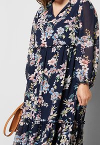 Námořnické květinové maxi šaty z lehkého materiálu s dlouhými rukávy, volánovými vrstvami a pastelovými květinovými vzory. Mají výstřih do V.