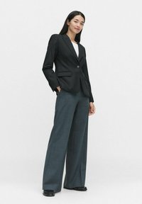 Svart skräddarsydd kavaj med en enda knapp, i kombination med vida mörkgrå byxor. Modellen bär en vit topp under. Slätt tyg.