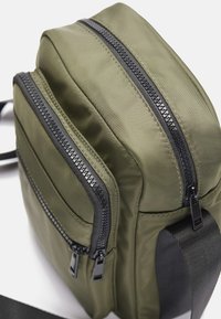 Zelena, pravokutna torba od glatkog materijala s više zip zatvarača i crnim detaljima. Uključuje podesivu naramenicu za nošenje.