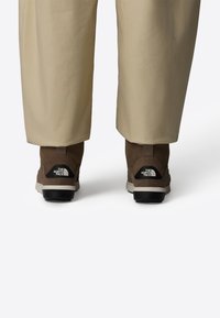 Stivali alla caviglia marroni con texture scamosciata, suole in gomma e accenti del logo neri. Abbinati a pantaloni beige a vestibilità ampia.
