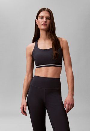 Kvinde med langt brunt hår iført sort sportstop og høj talje leggings, stående foran en ensfarvet grå baggrund.