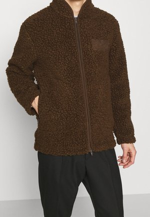 Homme portant une veste polaire zippée marron texturée avec poches latérales et pantalon noir, debout devant un fond blanc uni.