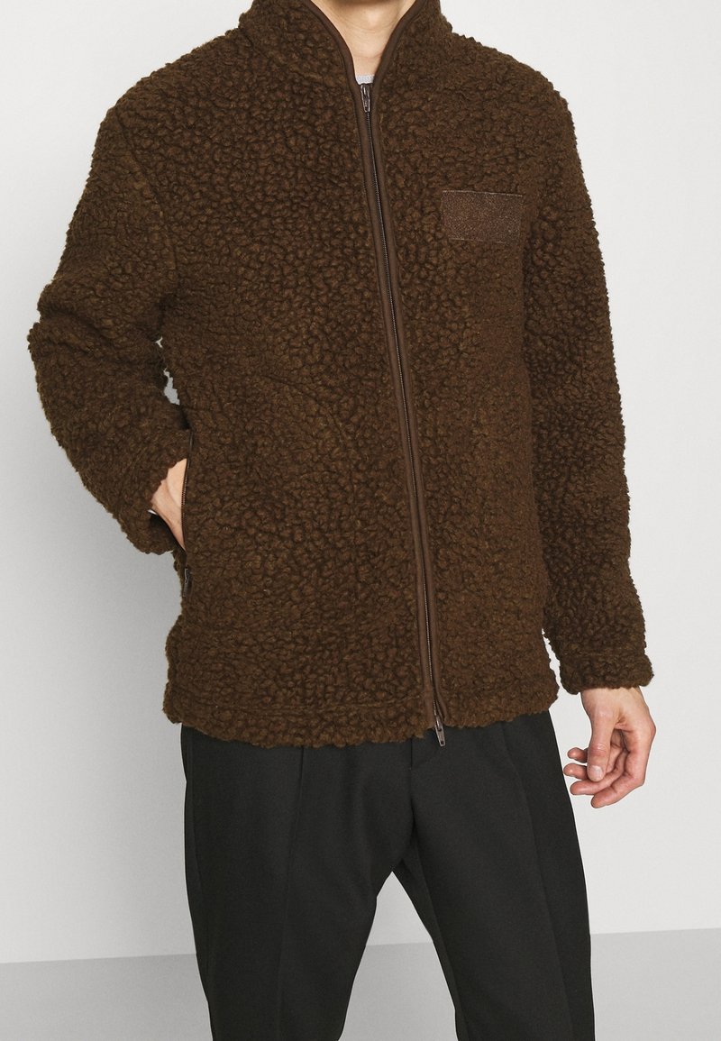 Homme portant une veste polaire zippée marron texturée avec poches latérales et pantalon noir, debout devant un fond blanc uni.