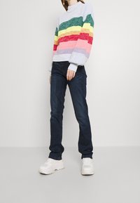 Suéter multicolorido con rayas horizontales en verde, amarillo, rojo y rosa, combinado con jeans azul oscuro y zapatillas blancas.