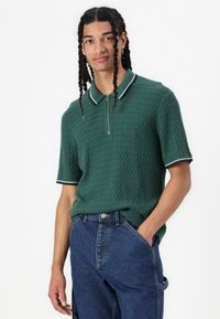 Polo shirt verde a maglia con maniche corte, caratterizzato da un motivo testurizzato e colletto e bordi delle maniche bianchi a contrasto. Indossato con jeans in denim blu.