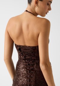 Robe en sequins marron avec un dos lacé et des accents en satin. Caractérisée par un col sans bretelles et une silhouette ajustée.