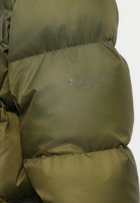 Sección de chaqueta acolchada verde oliva con tejido brillante y acolchado. Presenta un logo en relieve, costuras verticales y una textura suave.