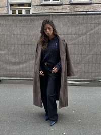 Gari pelēks pārmētelis pār tumši zilas krāsas diegu trikotāžas džemperi, kopā ar tumšām platajām džinsa biksēm un ziliem, asu purngalu augstpapēžu apaviem uz pelēka ietves.