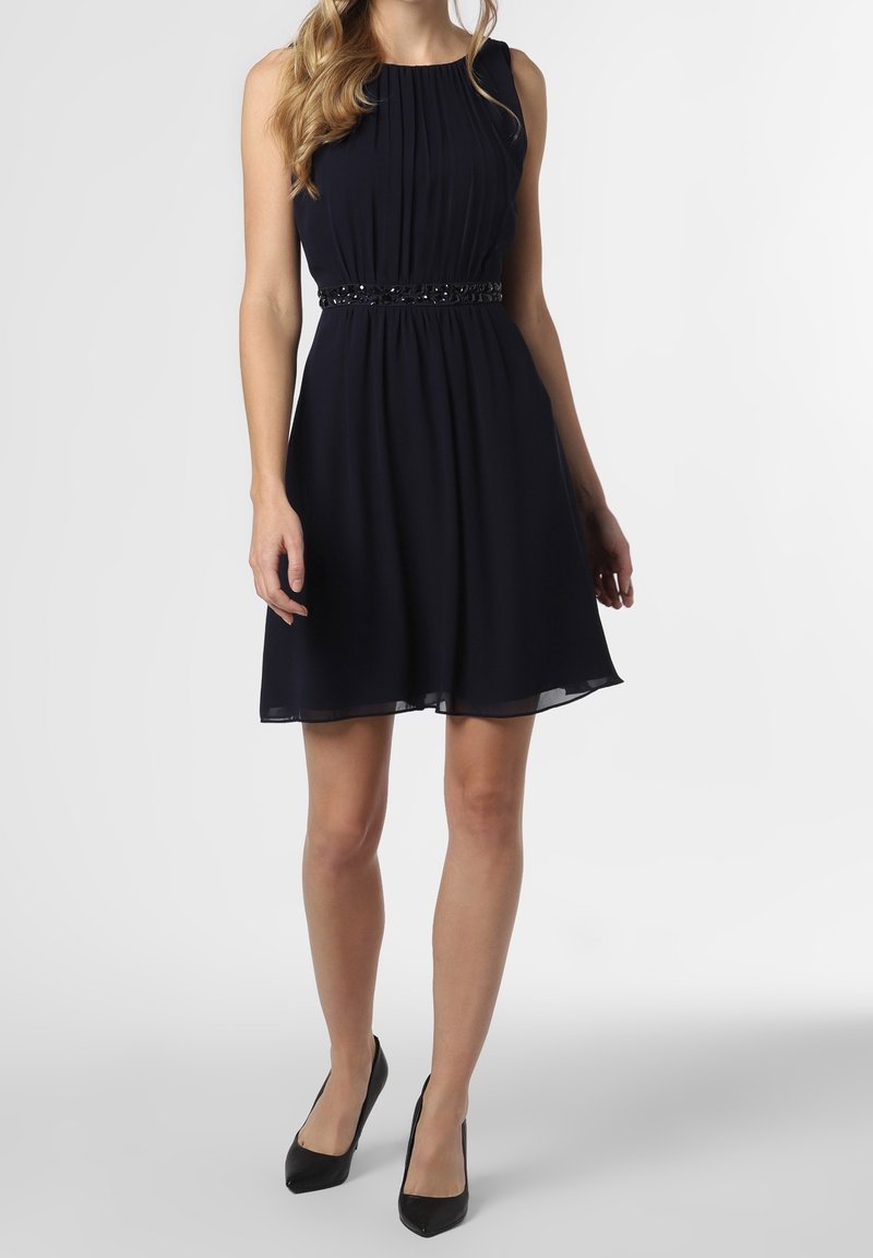 Kvinna i en ärmlös svart plisserad knälång klänning med prydd midja och svarta spetsiga klackskor på vit bakgrund.