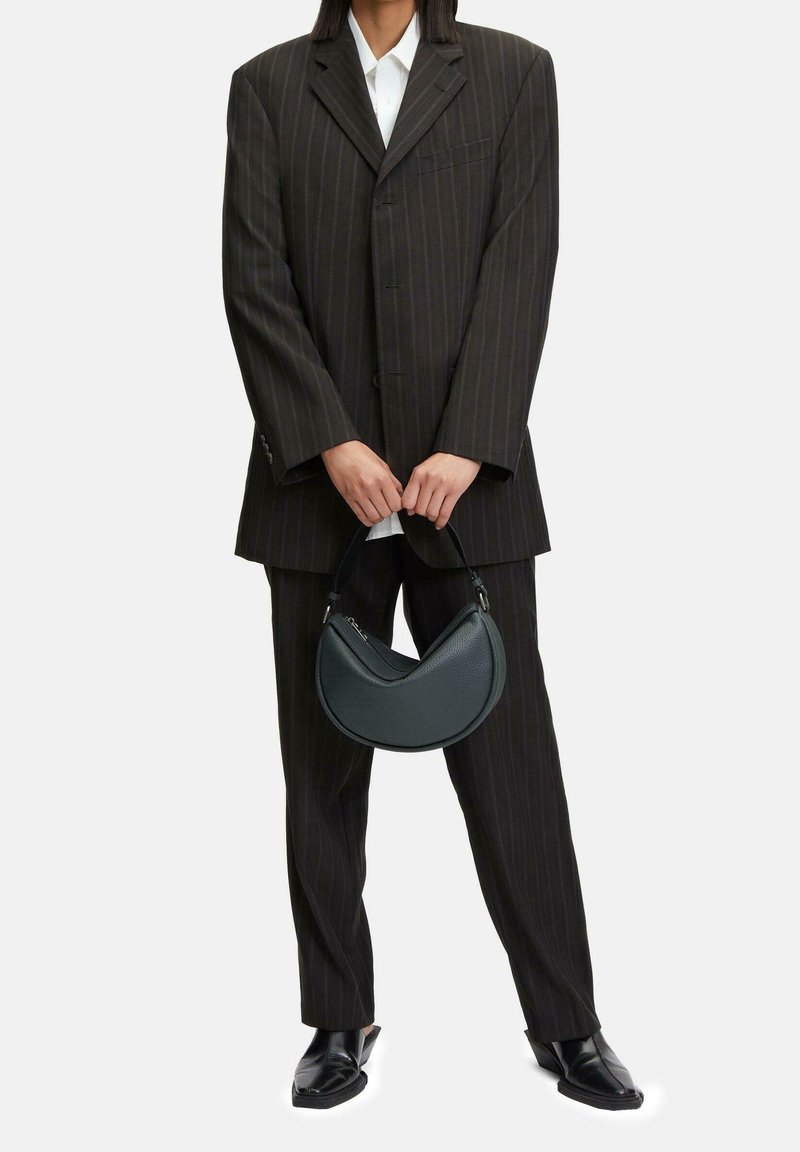 Personne portant un costume à fines rayures sombre oversize et des chaussures noires, tenant un petit sac à main noir en forme de croissant à deux mains.