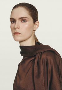 Bruine, gedrapeerde kledingstuk met een hoge kraag en een zachte textuur. De stof vertoont een subtiele glans en een vloeiende vormgeving met een gestrikte accent.