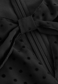 Tissu noir avec de petits pois en relief, présentant un nœud noué sur une fermeture éclair centrale fermée.
