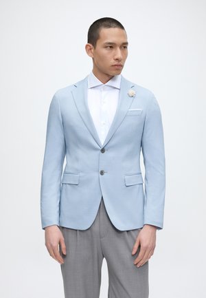 Homme portant un blazer bleu clair avec une épingle fleur, une chemise blanche et un pantalon gris, debout devant un fond blanc uni.