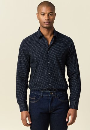 Camisa de botones azul marino con un pequeño patrón de lunares blancos, de manga larga y con cuello, combinada con jeans azul oscuro.