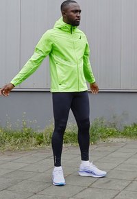 Giacca verde neon, leggera e con zip, abbinata a leggings neri. Scarpe sportive bianche e blu completano il look, posizionato su uno sfondo grigio.