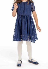 Ung flicka i en marinblå mönstrad klänning, vita strumpbyxor och marinblå Mary Jane-skor står mot en vit bakgrund.