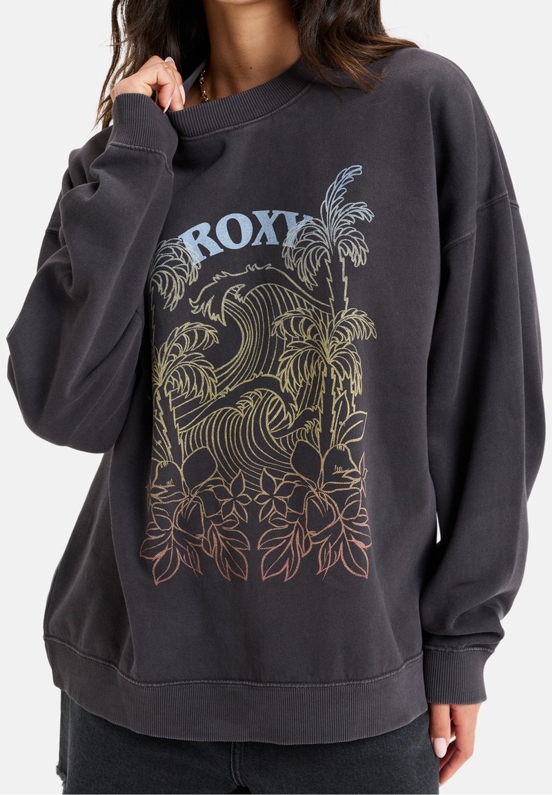 Femme portant un sweat-shirt oversize gris foncé avec un motif coloré de palmier, vague et fleur, et le texte "ROXY" à l'avant.