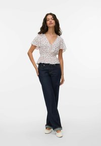 Kvinna med långt mörkt hår som bär en vit blommig knäppt blus, mörka uppvikta jeans och vita skor, stående mot en enkel vit bakgrund.