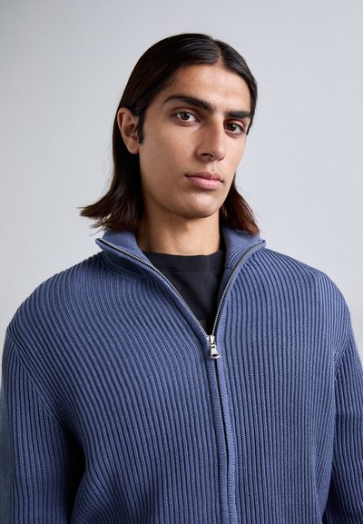 Μπλε πλεκτό ζακέτα με φερμουάρ, με ραβδωτή υφή, ψηλό γιακά και ασημένιο φερμουάρ. Φορεμένη πάνω από ένα μαύρο μπλουζάκι.