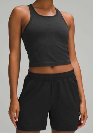 Femme portant un débardeur court noir sans manches et un short noir à taille élastique, debout devant un fond uni.