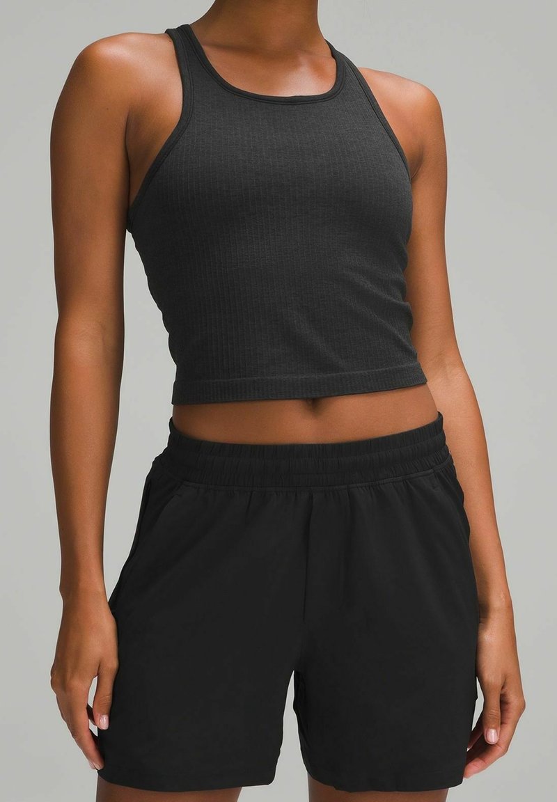 Femme portant un débardeur court noir sans manches et un short noir à taille élastique, debout devant un fond uni.