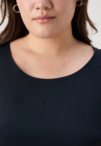 Top blu navy con texture, collo rotondo e tessuto liscio, che mette in mostra le spalle e la zona superiore del petto. Visibili orecchini a cerchio argentati.
