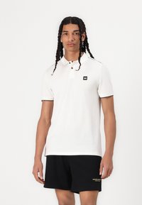 Hvit polo skjorte med svart logo på brystet, korte ermer og svart kant på kragen; brukt med svarte shorts med gul tekst.