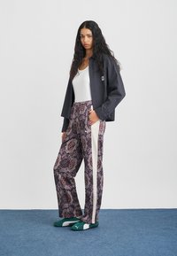 Μπλε πουκάμισο με καρό, λευκό αμάνικο τοπ, μοτίβο με μοβ παντελόνια με λευκές πλευρικές ρίγες και πράσινα παπούτσια τύπου σλάιντ. Το μοντέλο στέκεται μπροστά σε απλό φόντο.