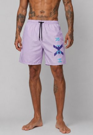 Uomo con tatuaggi che indossa pantaloncini da bagno viola chiaro con coulisse e scritte "R X H" blu e viola scuro sulla gamba destra, in piedi a piedi nudi.