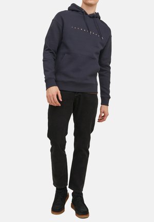 Marineblauer Hoodie mit einer vorderen Kängurutasche, verstellbarem Kapuzenzug und goldfarbenem Logodruck. Kombiniert mit schwarzen Hosen und dunklen Stiefeln. Material aus Baumwollmischung.