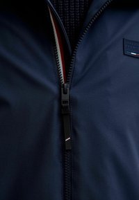 Marinejacke mit Reißverschluss vorne, aus einem strukturierten Stoff, schwarzer Reißverschlusszieher und einem Etikett mit rotem, weißem und blauem Akzent.