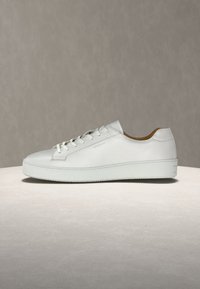 Balti odinė žemo profilio sportbačių pora su raišteliais, storu padu ir ruda vidine oda, eksponuojama ant neutralaus paviršiaus prieš pilką foną.