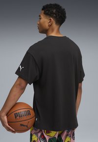 Czarna koszulka krótki rękaw z okrągłym dekoltem i subtelnym logo Puma na rękawie. Model trzyma w rękach piłkę do koszykówki, nosi spodenki w wzory.