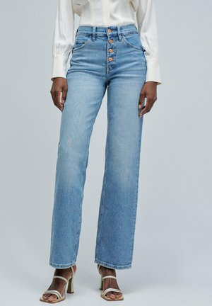 Mujer llevando jeans rectos de tiro alto en un azul claro con botones delanteros y sandalias de tacón blanco con tiras, contra un fondo liso.