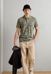 Kurzärmliges grünes Poloshirt mit Blumenmuster, kombiniert mit beigen Hosen und einer schwarzen Lederjacke, vor einem neutralen Hintergrund.