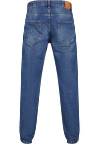 Jeans din denim de culoare albastru mediu, cu picior tapered, două buzunare la spate și o etichetă din piele maro la talie. Material moale și texturat.